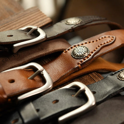 Three stacked leather belts in black, tan, and dark brown with silver buckles and decorative metal concho studs on wood surface.