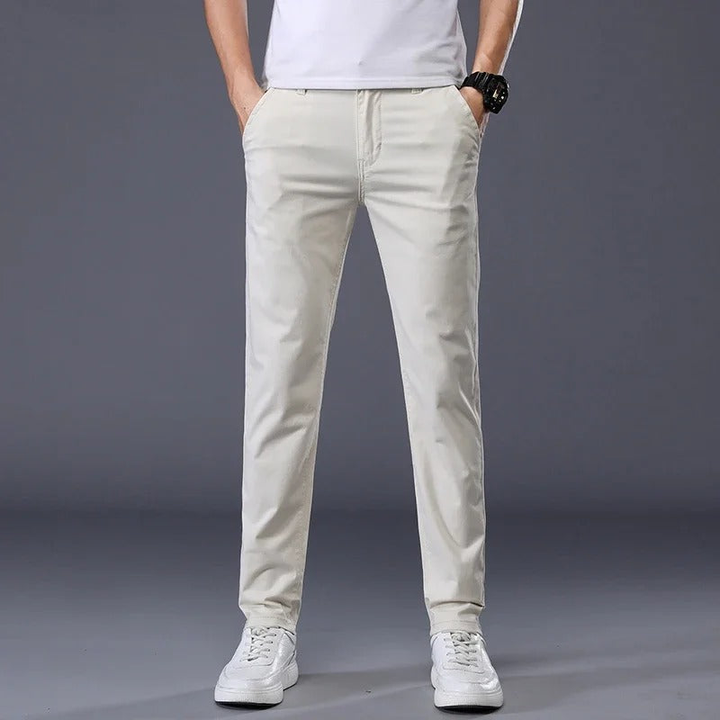 Man wearing slim fit beige cotton dress pants with white t-shirt and sneakers on gray background