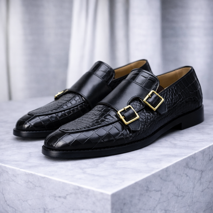 Black croc-embossed leather double monk strap loafers with gold buckles displayed on marble surface.