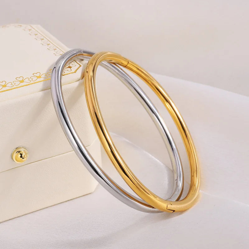 Two smooth polished bangles in gold and silver tones leaning against a white jewelry box on white fabric.
