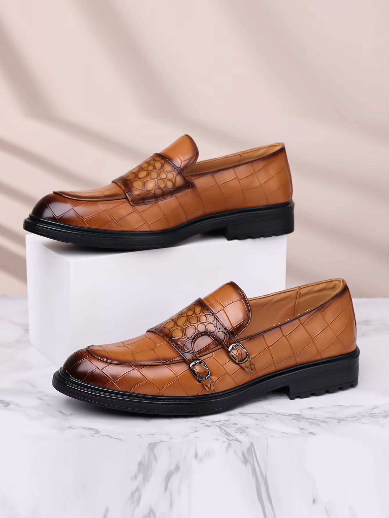 Tan croc-embossed leather men's loafers with double monk strap buckles and black rubber sole on marble surface.