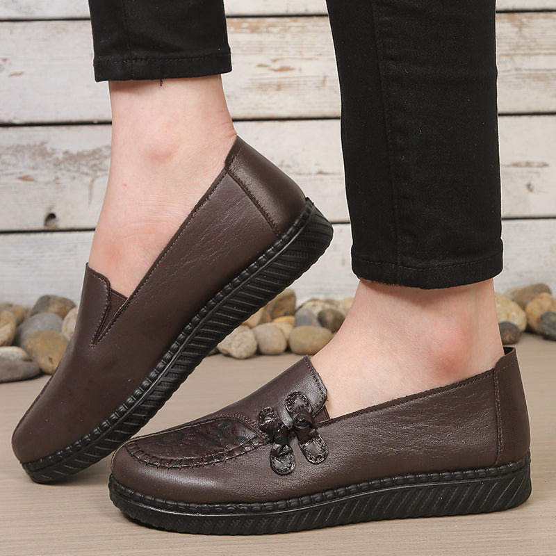 Brown leather women's slip-on loafers with decorative stitching and textured black platform sole, worn with black pants.