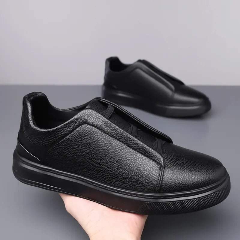 Black leather slip-on sneakers with contrast trim and thick sole, held in hand with matching pair in background