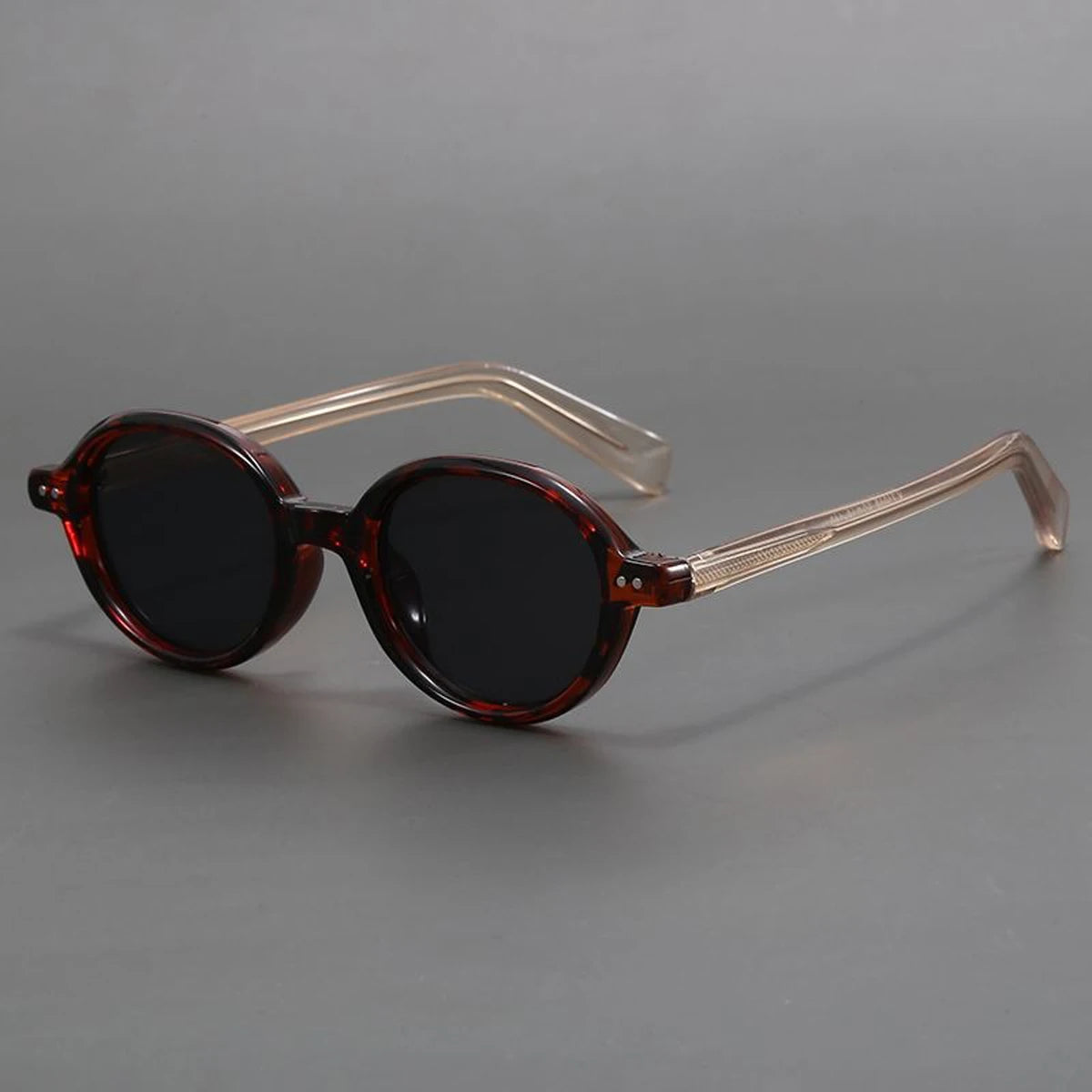 Round sunglasses with dark tortoiseshell frames, black lenses, and transparent champagne-colored temples on gray background.
