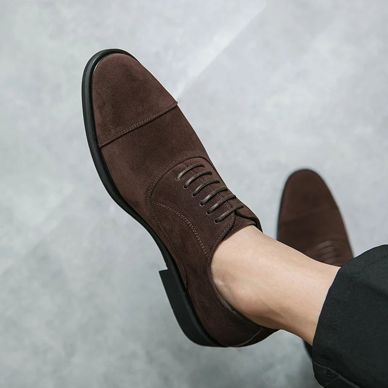 Brown suede cap-toe Oxford dress shoes with lace-up closure and black sole, worn with dark trousers