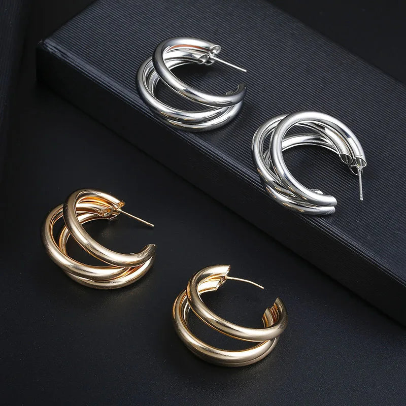 Women's gold triple-hoop twist earrings with snap closure, shown alongside matching silver pair on dark background.