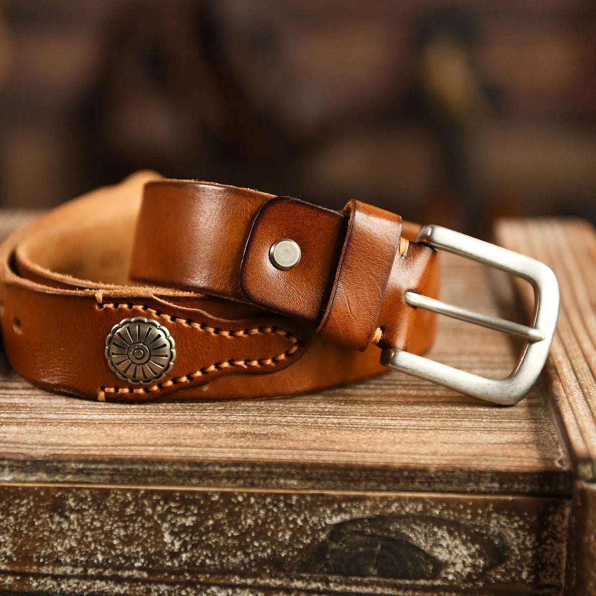 Hand-stitched brown leather belt with silver metal concho medallions and brushed steel buckle on rustic wooden background