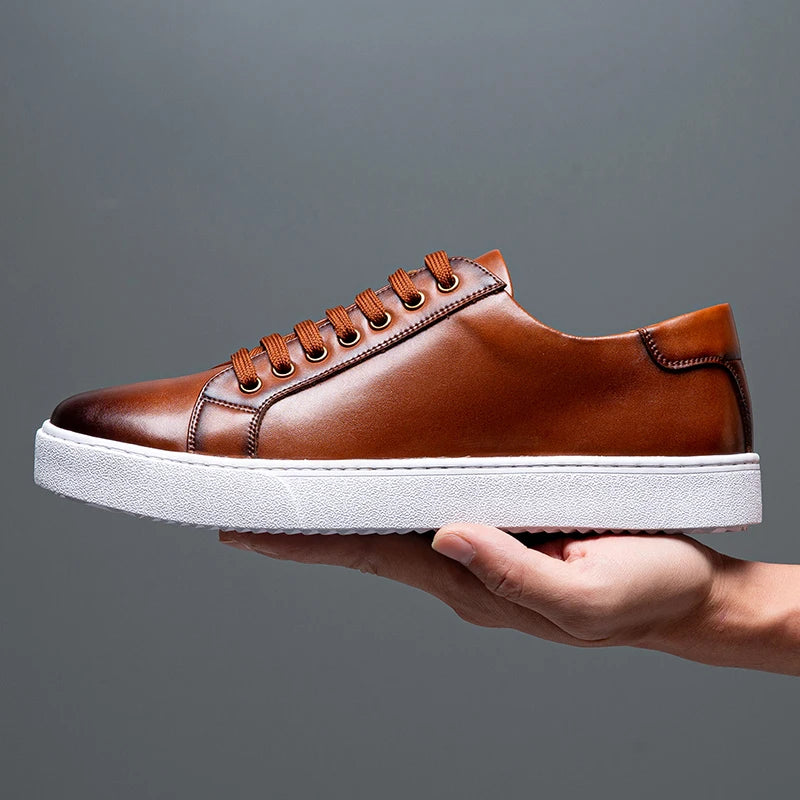 Brown leather lace-up sneakers for men with white rubber sole, held in hand against gray background