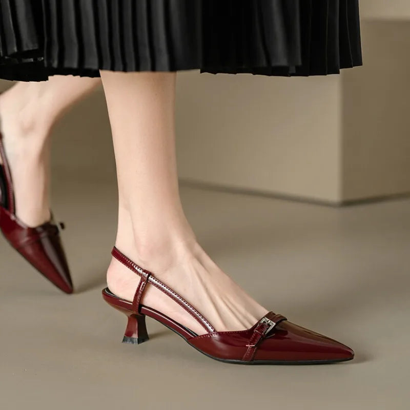 Burgundy patent leather pointed-toe slingback pumps with kitten heel and metal buckle detail, worn with black pleated skirt