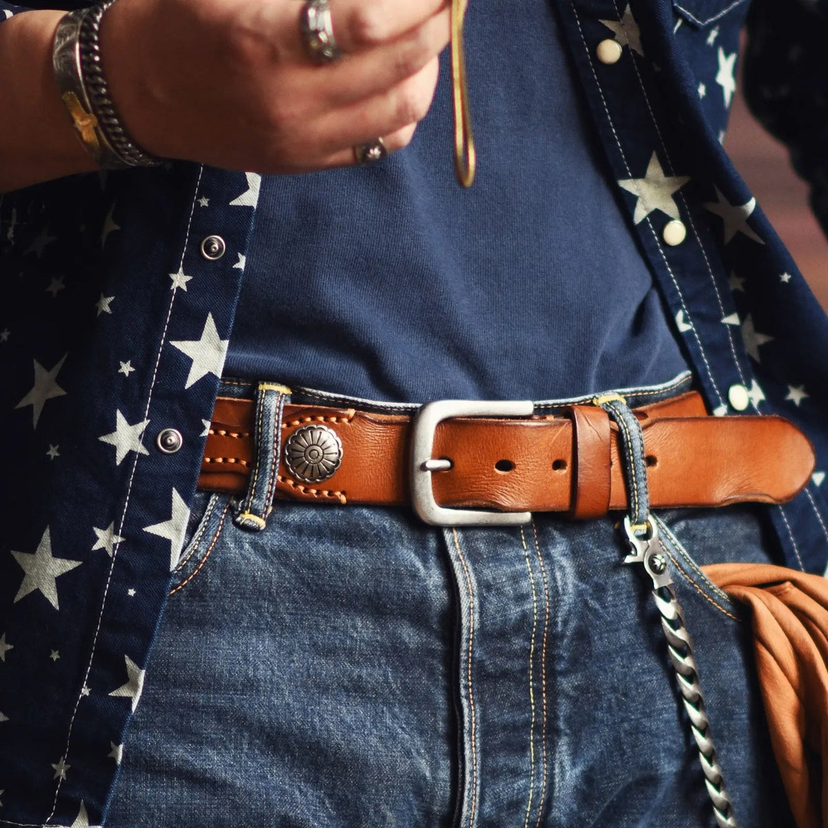 Brown leather belt with silver concho and square buckle, hand-stitched detailing, worn with denim jeans and star-print shirt.
