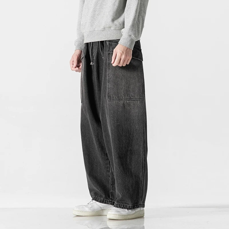 Man wearing dark gray wide-leg denim jeans with large front pocket, paired with gray sweatshirt and white sneakers