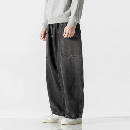Man wearing dark gray wide-leg denim jeans with large front pocket, paired with gray sweatshirt and white sneakers
