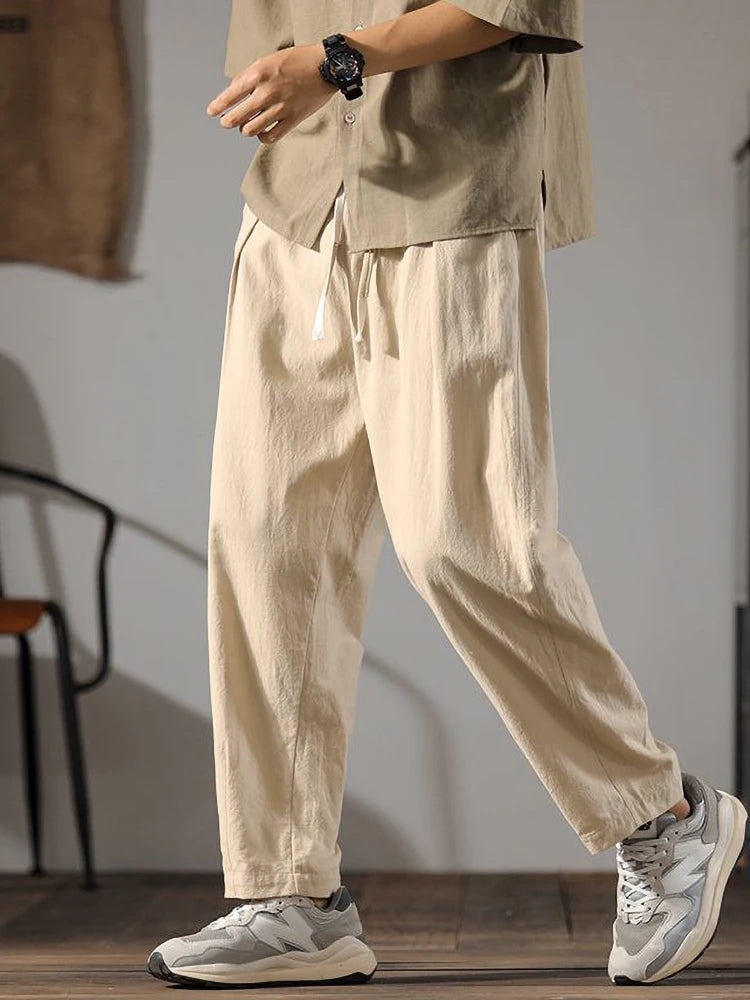 Man wearing beige wide-leg drawstring linen pants with matching shirt and gray sneakers, casual relaxed fit style.