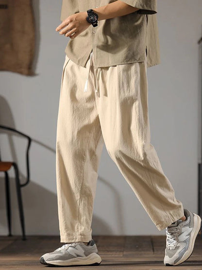 Man wearing beige wide-leg drawstring linen pants with matching shirt and gray sneakers, casual relaxed fit style.