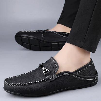 Black men's leather slip-on loafers with white stitching, metal buckle detail, and rubber sole, shown on feet with black trousers.
