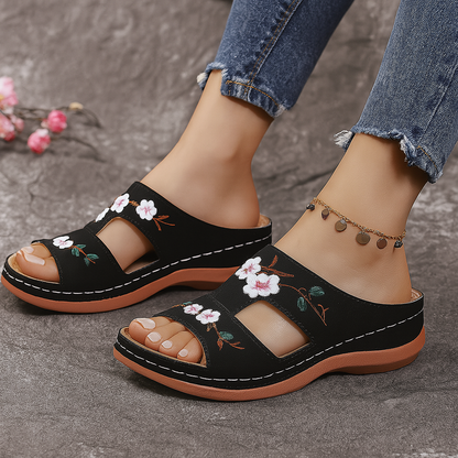 Black platform slide sandals with white floral embroidery, open toe, tan sole, worn with distressed jeans and gold anklet.