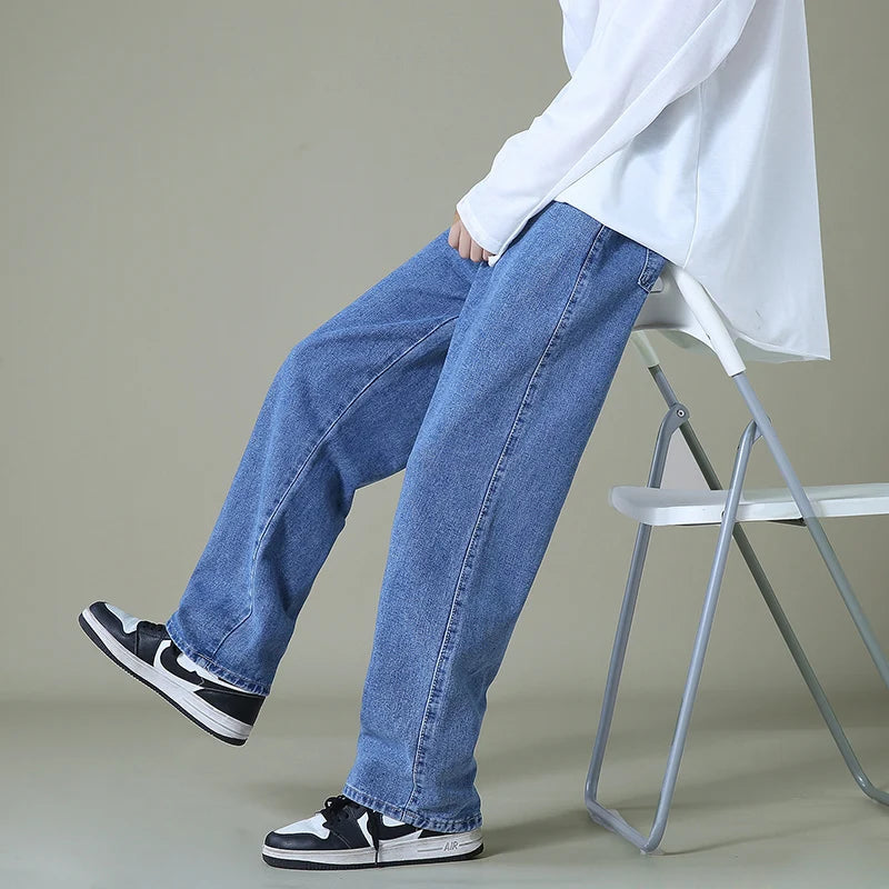 Man sitting on white chair wearing relaxed-fit light blue denim jeans, white long-sleeve shirt, and black Nike sneakers.