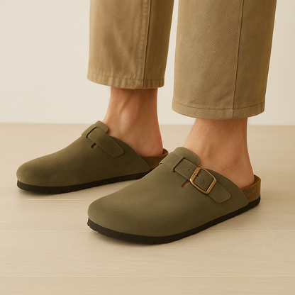 Men's olive green suede slip-on clogs with gold buckle detail and dark sole, styled with khaki trousers.