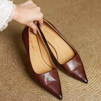 Women's burgundy leather pointed-toe pumps held by hand, tan interior lining, displayed on woven surface.