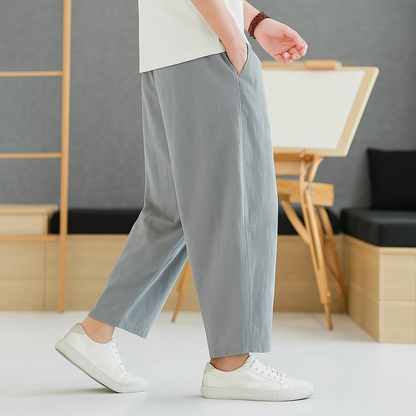 Man wearing light gray tapered linen-cotton pants with white sneakers and a white t-shirt in a minimalist setting.