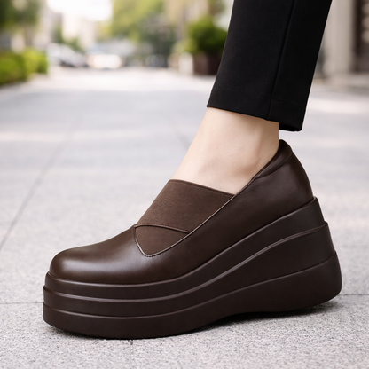 Women's brown genuine leather orthopedic platform slip-on shoes with thick stacked sole, worn with black pants outdoors.