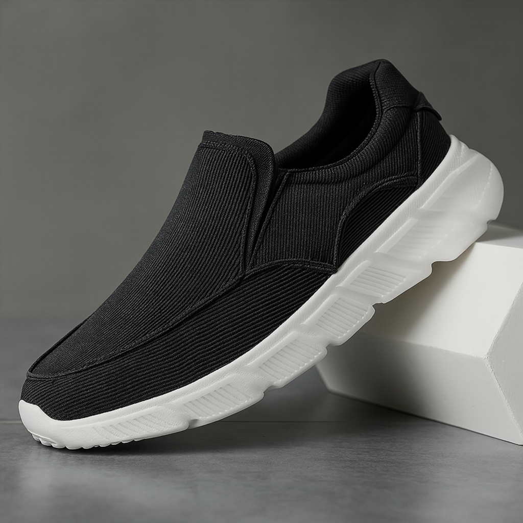 Black corduroy slip-on sneakers with white chunky sole, displayed against a gray background on white blocks.