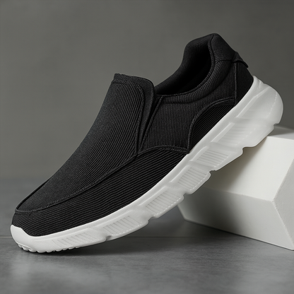 Black corduroy slip-on sneakers with white chunky sole, displayed against a gray background on white blocks.