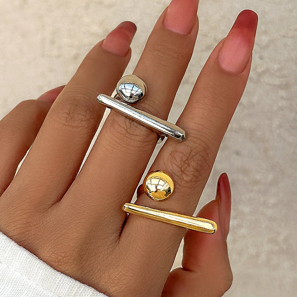 Woman's hand wearing two adjustable open bar and ball rings in silver and gold tones with pink ombre nails