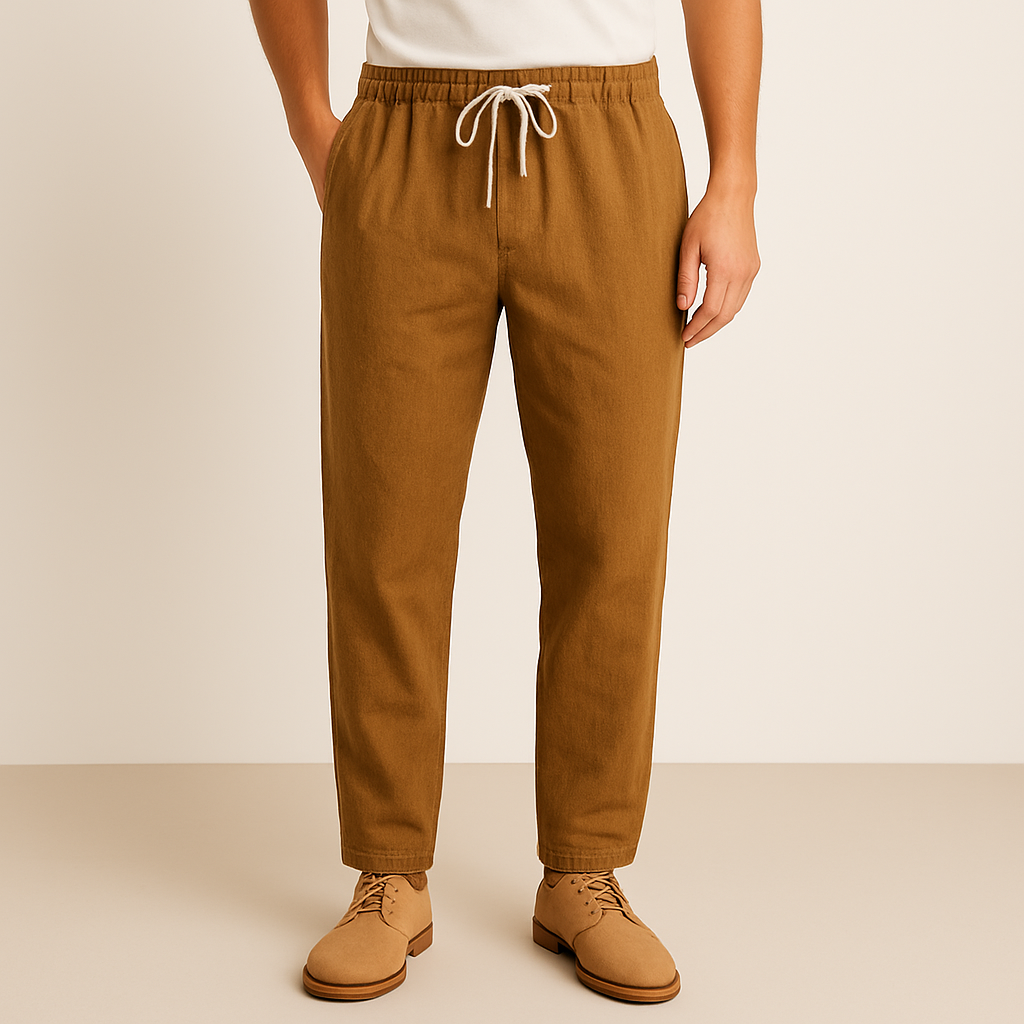 Men's tan linen-cotton drawstring pants with elastic waistband, tapered fit, worn with white tee and tan oxford shoes.