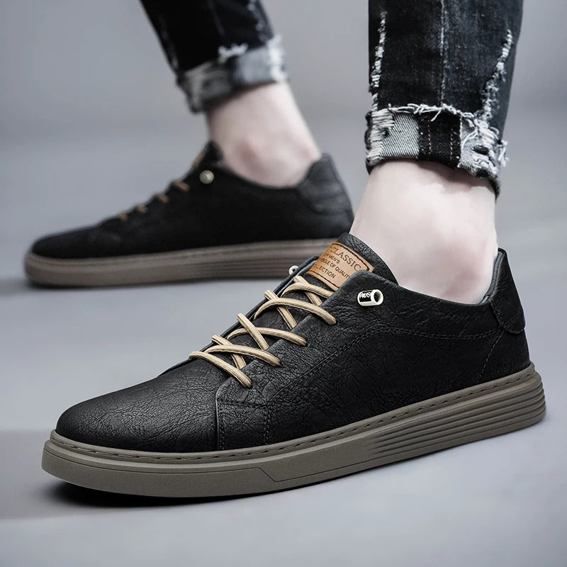 Black leather low-top men's sneakers with tan laces and brown sole, styled with ripped jeans on gray background.