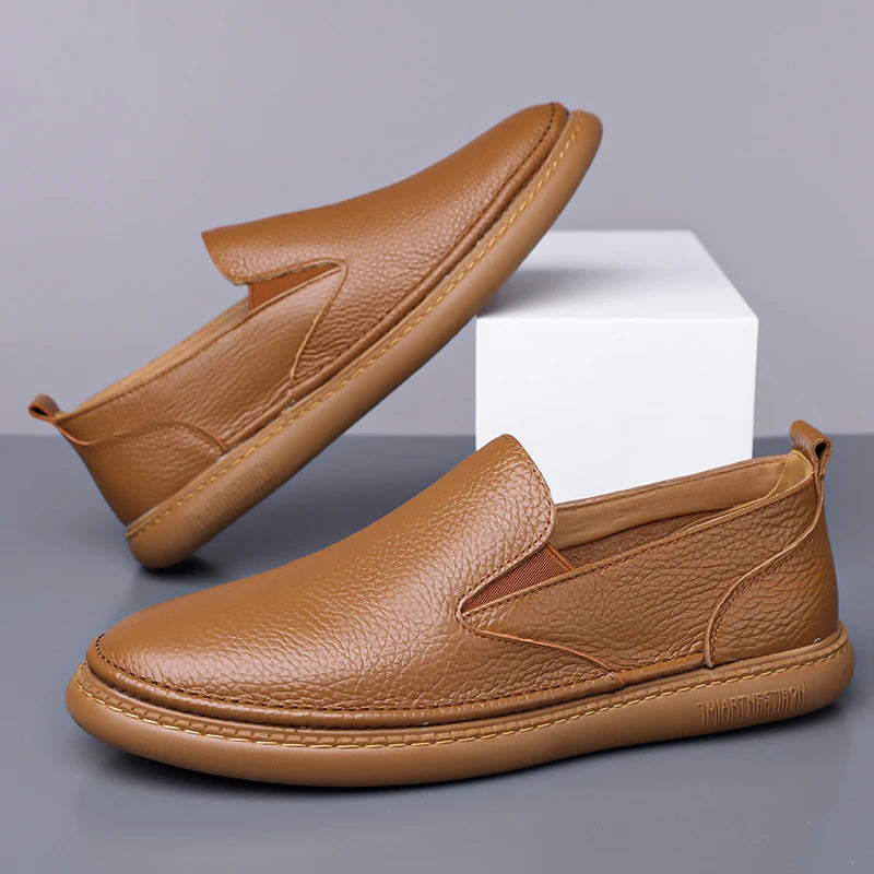 Men's tan brown leather slip-on loafers with textured grain finish and cushioned sole displayed on white platform
