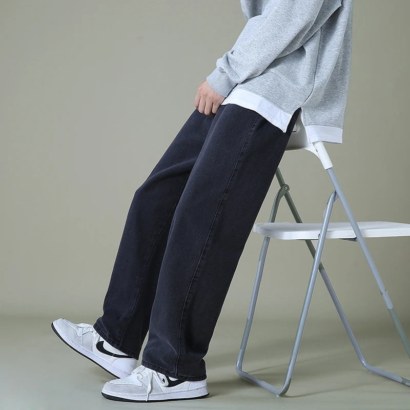 Man sitting on white folding chair wearing dark navy relaxed-fit denim jeans, gray sweatshirt, and white sneakers.