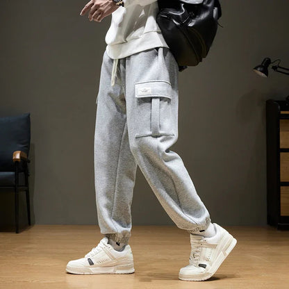 Men's gray cargo joggers with side pockets, drawstring waist, elastic cuffs, paired with white sneakers and backpack.