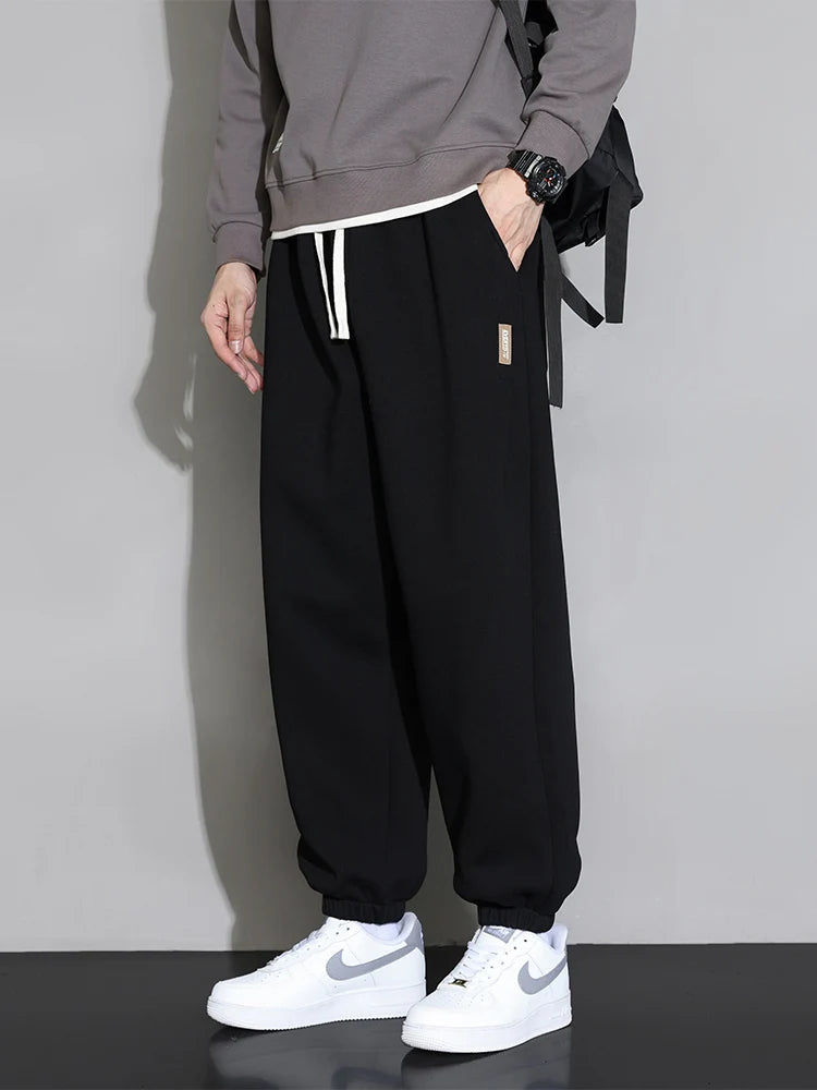 Men's black relaxed fit cotton joggers with white drawstring, worn with gray sweatshirt and white Nike sneakers