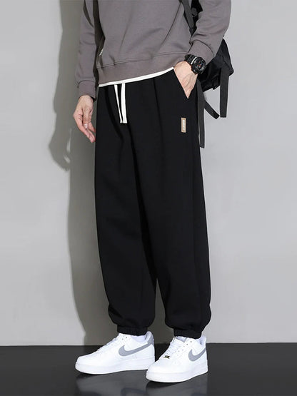 Men's black relaxed fit cotton joggers with white drawstring, worn with gray sweatshirt and white Nike sneakers