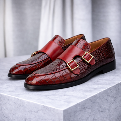 Burgundy croc-embossed leather double monk strap loafers with gold buckles and black sole on marble surface