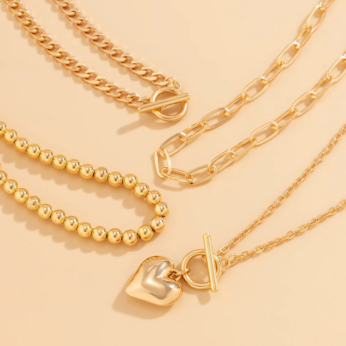 Gold layered necklace set featuring curb chain, bead chain, paperclip chain, and delicate chain with heart pendant toggle clasps