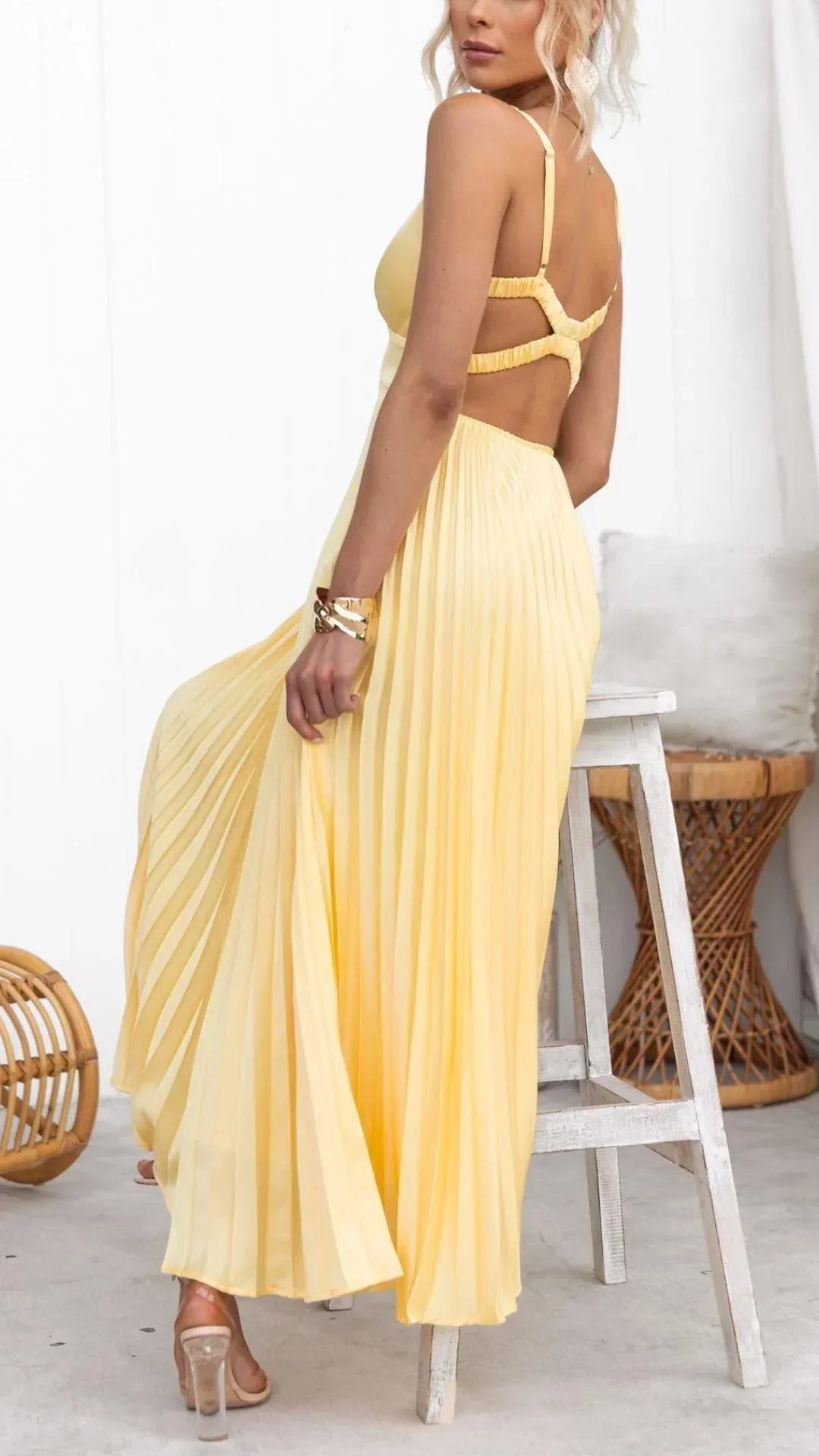 Yellow pleated maxi dress with open back strappy detail, V-neck, and flowing skirt, paired with clear heeled sandals.