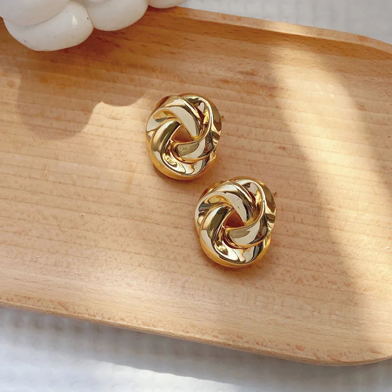 Gold twisted knot stud earrings with snap closure displayed on wooden tray, polished metallic finish