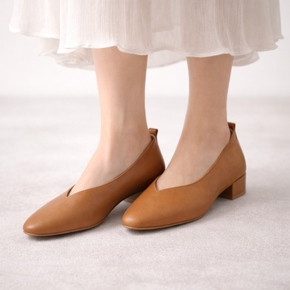 Women's tan leather block heel pumps with V-cut vamp, low stacked heel, worn with white midi dress on grey background.