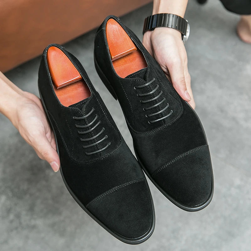 Black suede cap-toe Oxford dress shoes with lace-up closure and cedar shoe trees, held by a man wearing a watch.