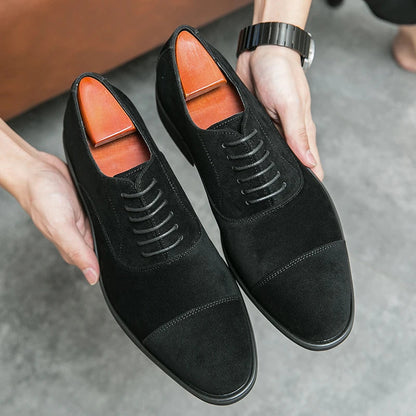 Black suede cap-toe Oxford dress shoes with lace-up closure and cedar shoe trees, held by a man wearing a watch.