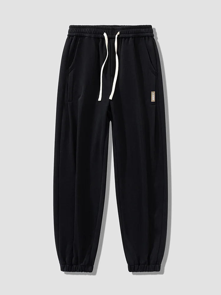 Black men's relaxed fit cotton jogger pants with white drawstring waist, elastic cuffs, side pockets, and small label patch.