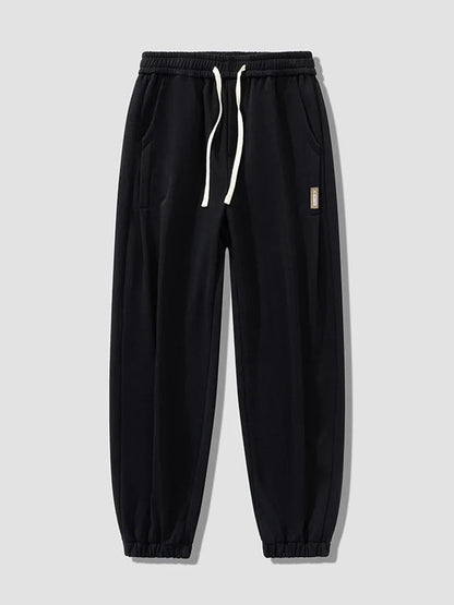 Black men's relaxed fit cotton jogger pants with white drawstring waist, elastic cuffs, side pockets, and small label patch.