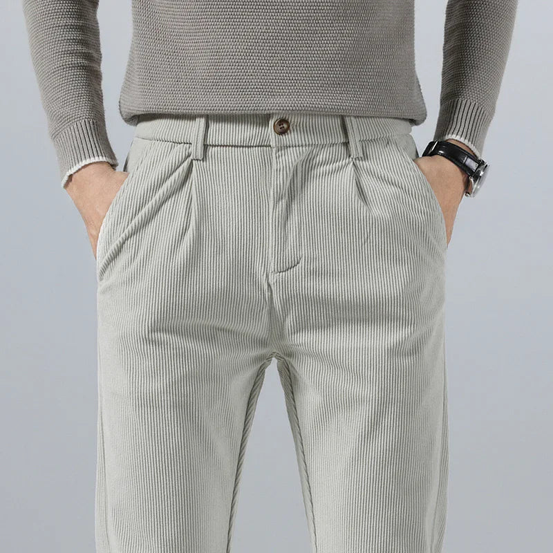 Light gray men's slim fit corduroy pants with pleated front, side pockets, and brown button closure, paired with gray sweater.