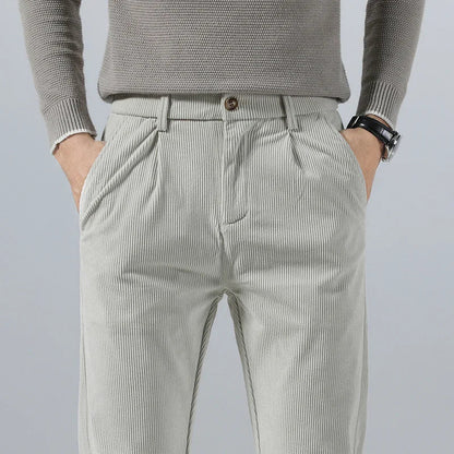 Light gray men's slim fit corduroy pants with pleated front, side pockets, and brown button closure, paired with gray sweater.