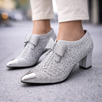 Silver rhinestone pointed-toe block heel booties with decorative bow and metallic cap toe, worn with white pants.