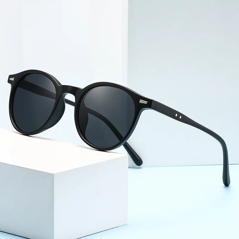 Black round acetate sunglasses with dark lenses and slim temples displayed on a white geometric block
