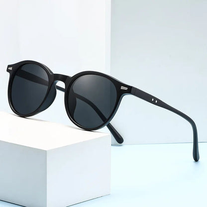 Black round acetate sunglasses with dark lenses and slim temples displayed on a white geometric block