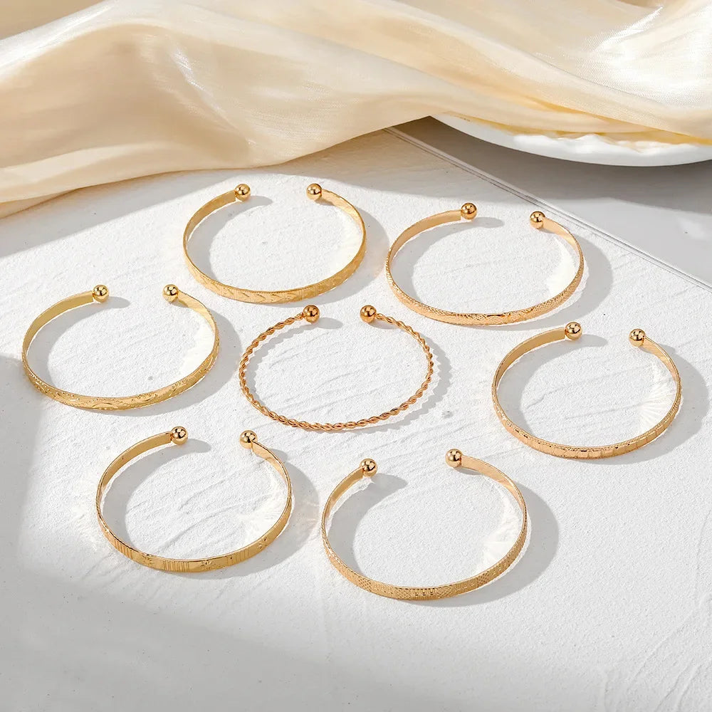 Seven gold open cuff bangle bracelets with ball ends, featuring etched patterns and twisted designs, displayed on white surface.